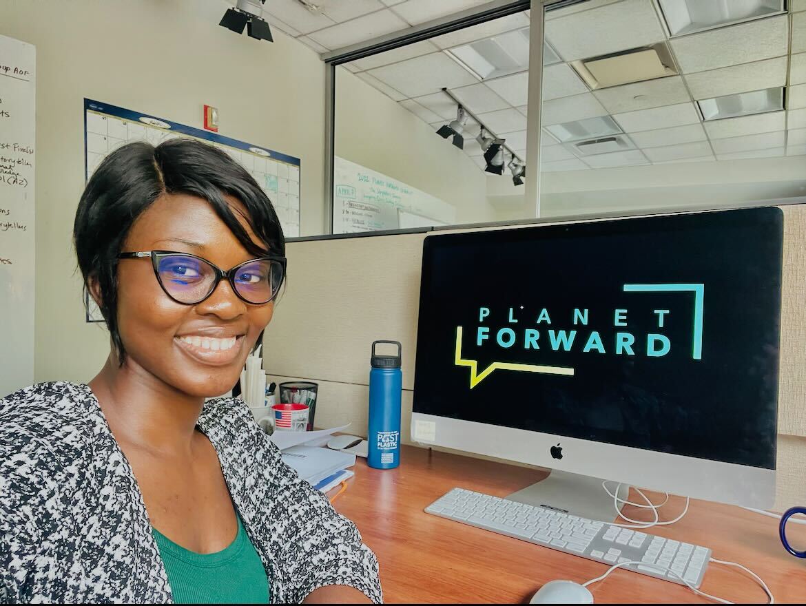 Beverly smiles at the camera while sitting at a computer with the Planet Forward logo prominently displayed. Planet Forward is a non-profit that specializes in environmental journalism.